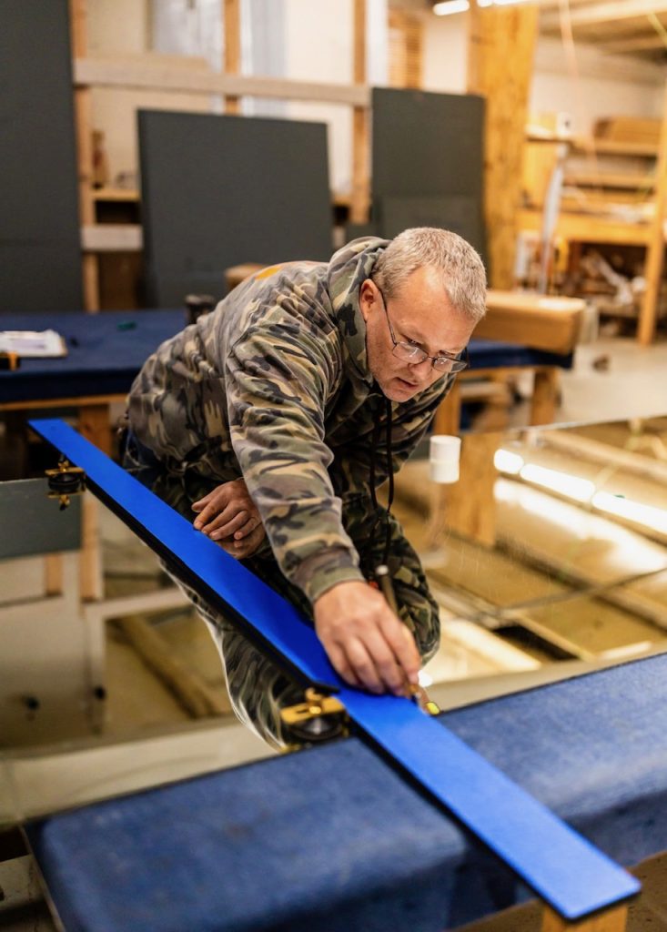 A craftsman wearing eyeglasses and a camouflage jacket leaning over a workbench to make precise measurements along a bright blue straight edge.