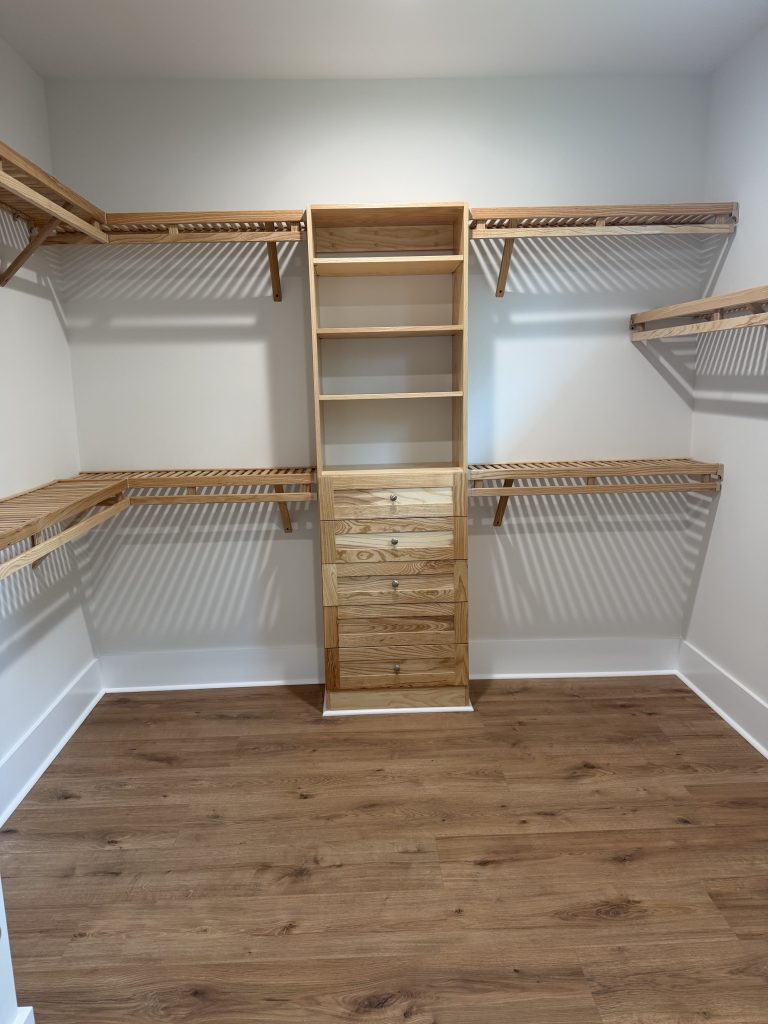 A sideways wide-angle shot of a custom light-wood walk-in closet storage unit with open shelving cubbies, five built-in drawers, and slatted hanging racks mounted on white walls above light wood flooring.