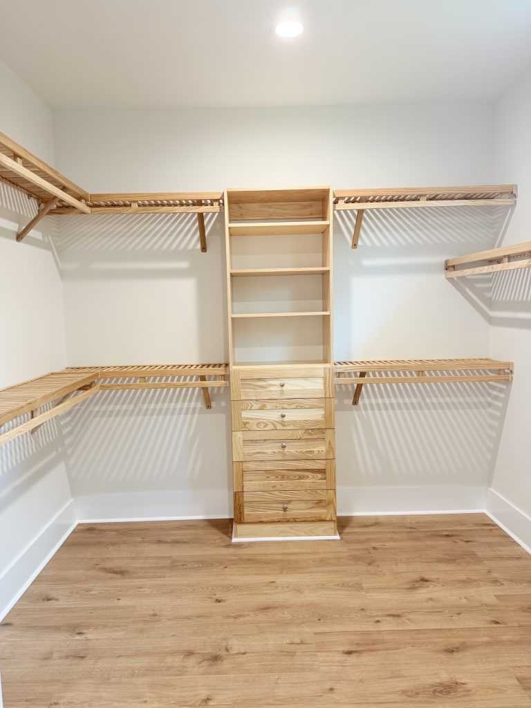 A sideways wide-angle shot of the light-wood closet shelving system, illuminated by a bright overhead light that highlights the wooden drawers, open shelves, and slatted clothing racks against white walls.