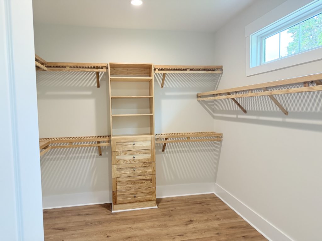 An upright view of an empty walk-in closet featuring a custom light-wood organizer tower with open shelves on top and five drawers below. Multi-level slatted racks with hanging rods flank the tower on white walls, set over light hardwood floors next to a small, high window.